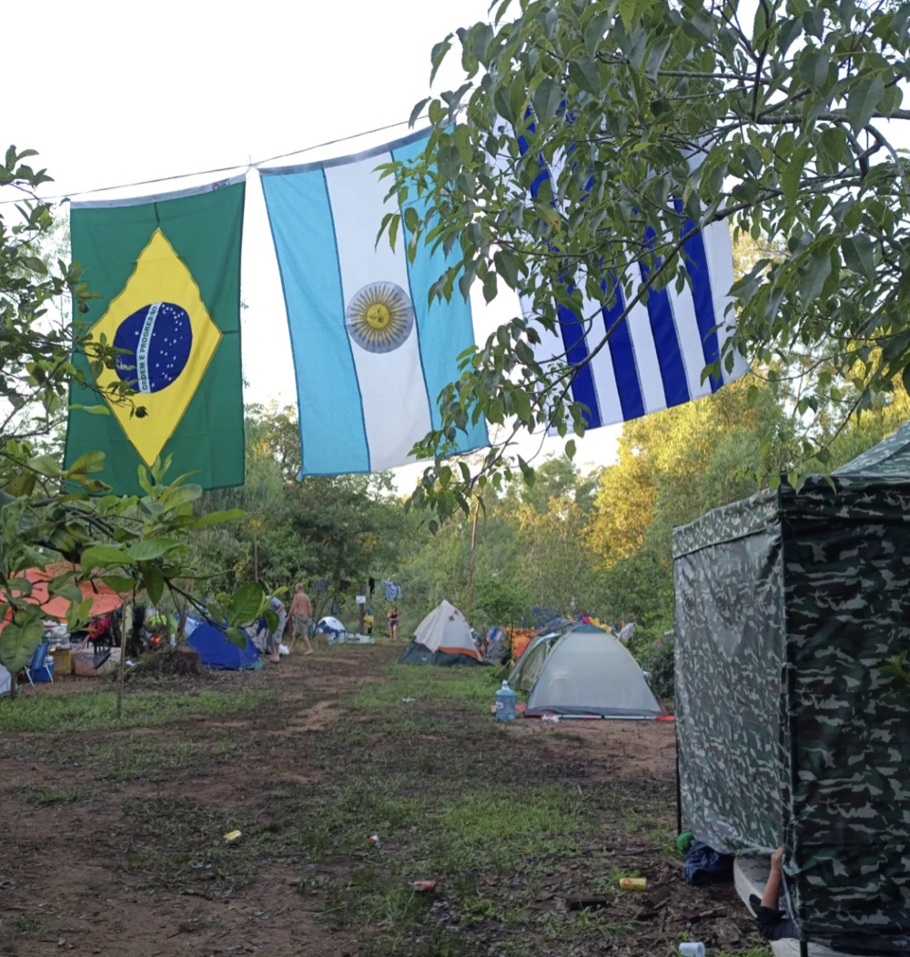 Acampamento com as bandeiras de Brasil, Argentina e Uruguai estendidas entre as árvores